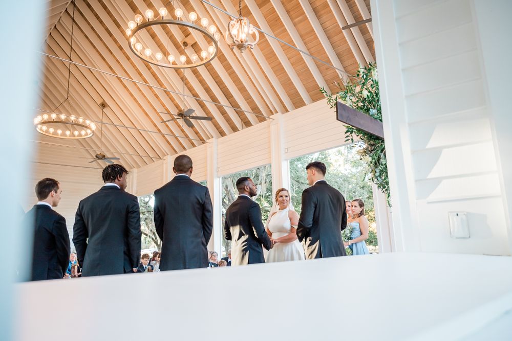 Groomsmen in black suits stand together under a wooden chapel ceiling with pendant lighting.