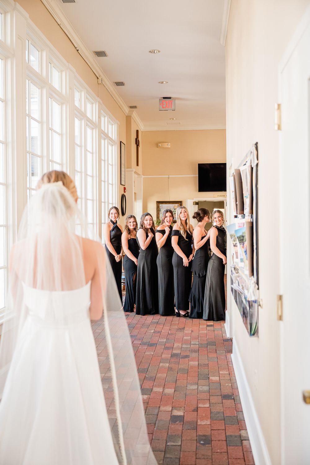 Bridesmaids in black dresses react to bride's first look reveal in sunlit hallway.