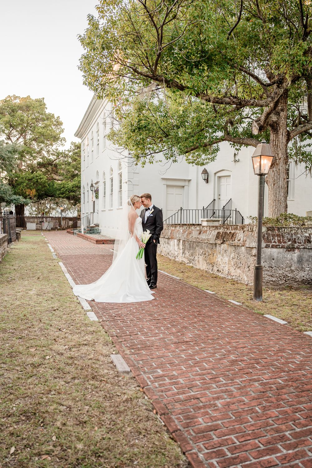 Elegant couple posing on brick pathway in front of historic white building with trees.