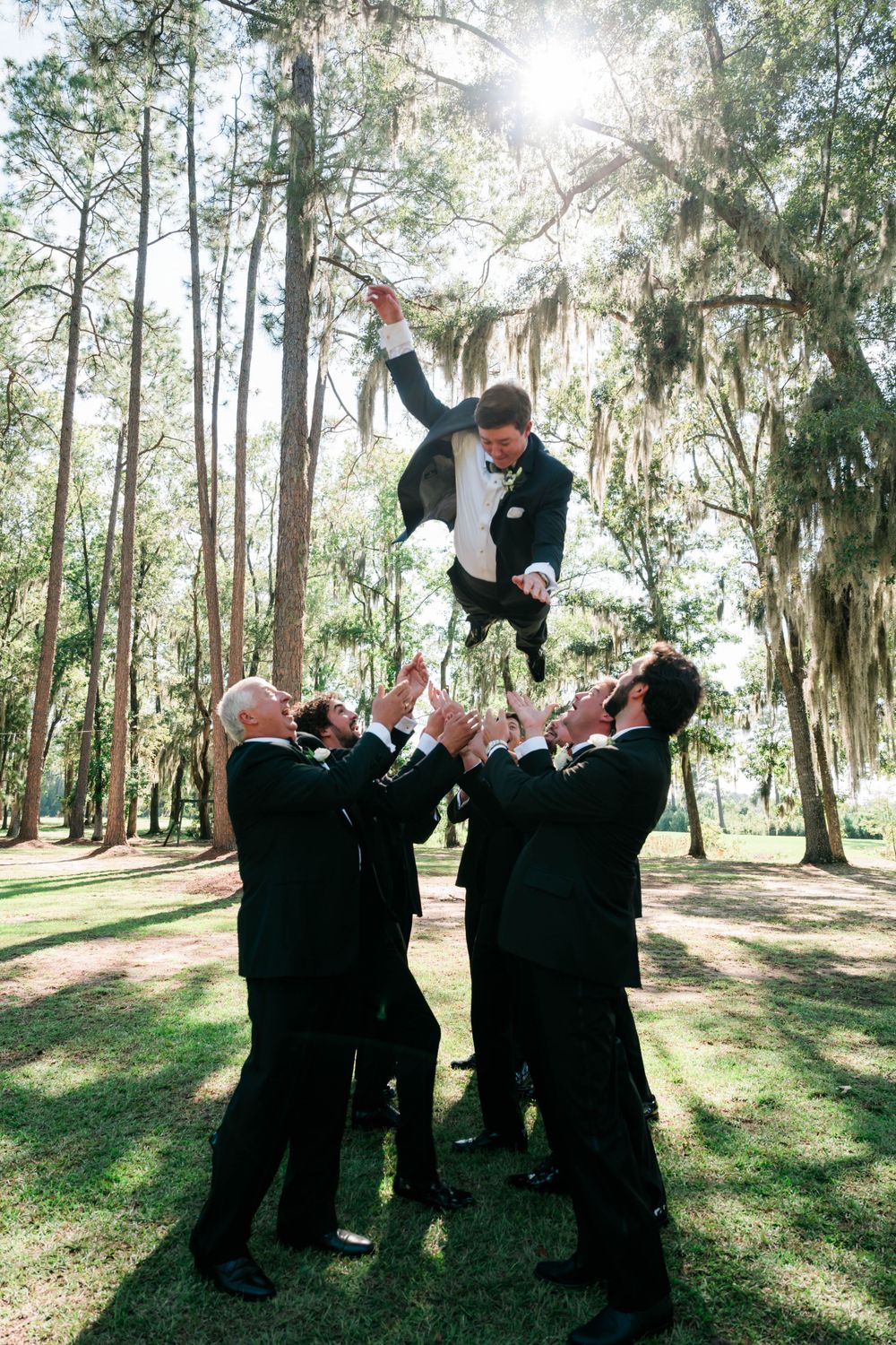Celebratory group photo with groomsmen tossing groom in wooded outdoor setting.