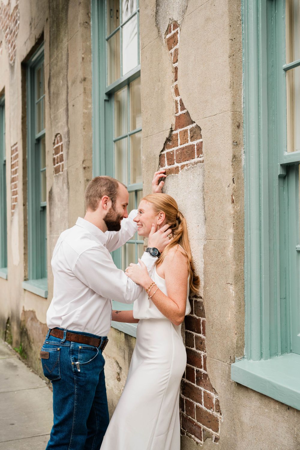 Intimate portraits of couple embracing against rustic brick wall with turquoise windows.
