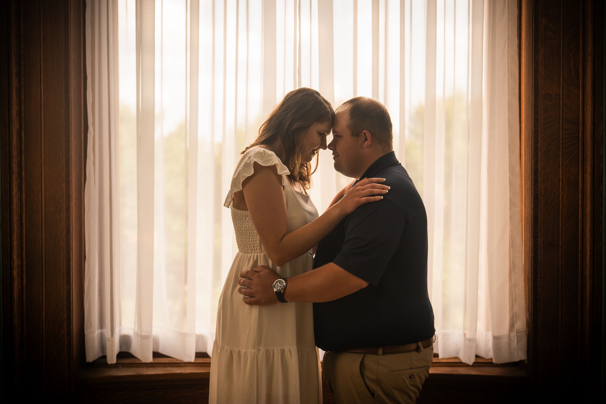 Couple sharing an intimate moment together, standing close by a large window with soft curtain light.