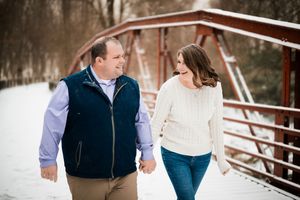 Couple holding hands on a snowy bridge in winter, wearing casual sweaters and smiling at each other.