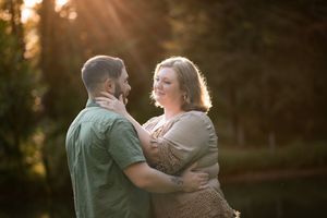 Couple embracing outdoors at golden hour sunset with warm sunlight glowing through trees in background.