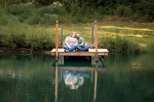 Couple sitting together on a wooden dock over a calm reflective lake surrounded by lush green trees.