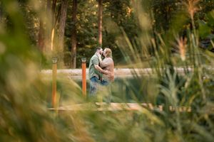 Couple sharing a romantic kiss on a dock surrounded by lush green trees and tall grass in a serene outdoor setting.