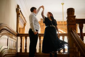 Couple dancing on ornate wooden staircase, man spinning woman in black dress inside elegant historic building.