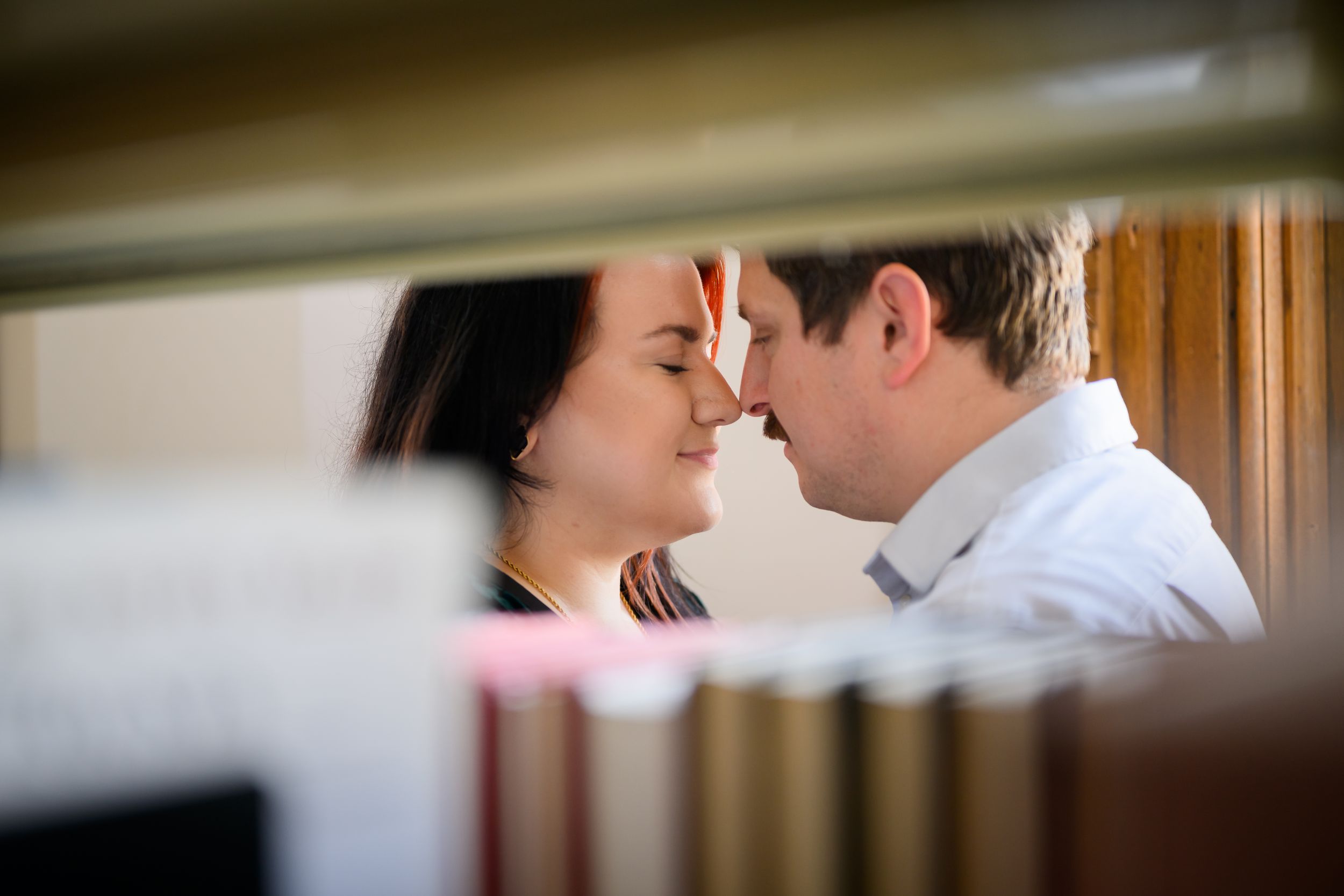 Romantic couple sharing an intimate moment, touching foreheads tenderly between library bookshelves.
