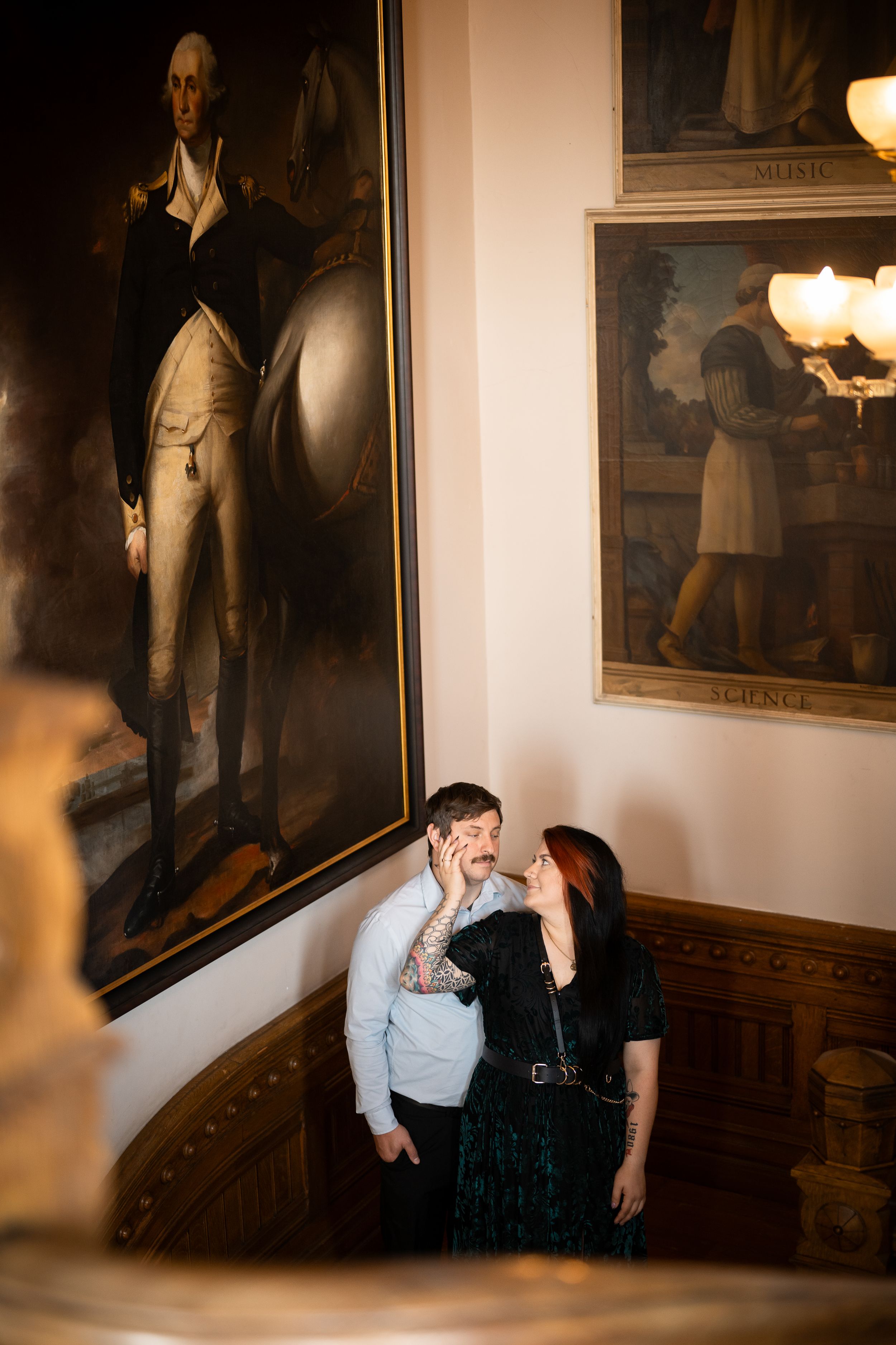 Couple shares romantic moment on grand staircase beside large George Washington portrait in historic building.