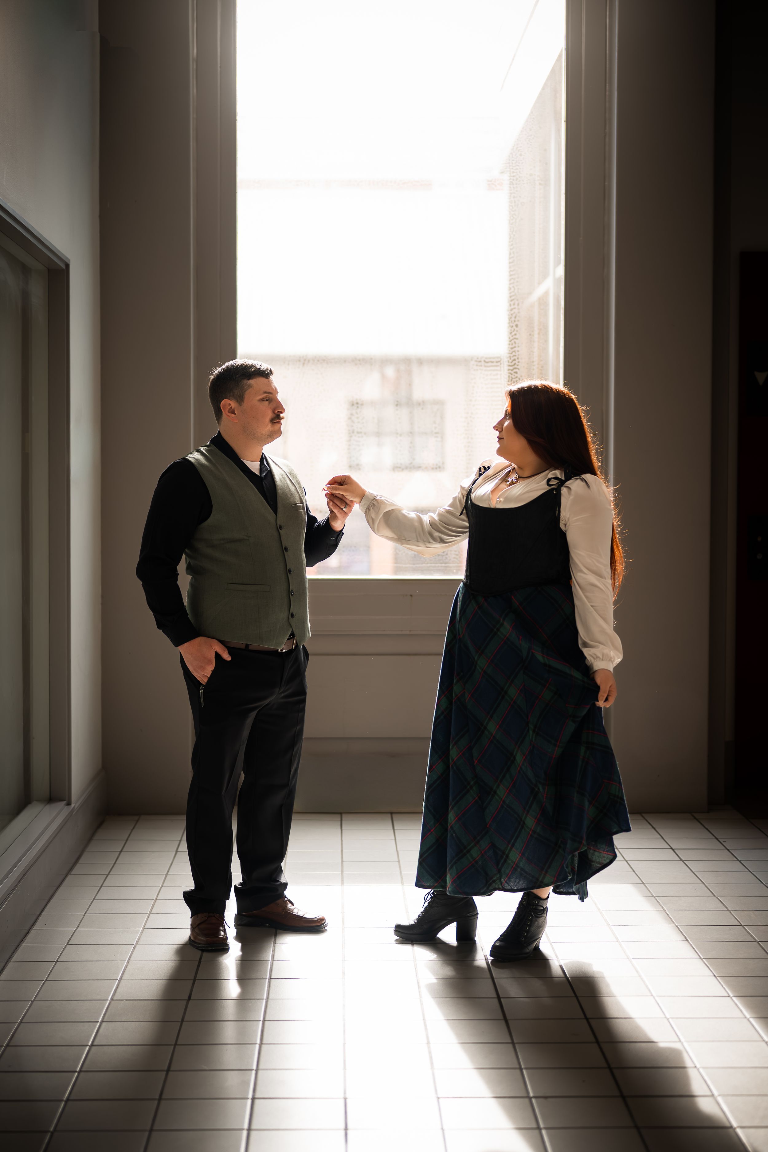 A couple holds hands and dances in a sunlit hallway, the woman in a plaid dress and the man in a vest.