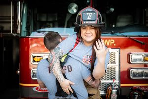 Woman in firefighter helmet smiling in front of red fire truck at station.