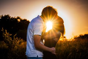 Couple silhouetted against golden sunset in a field, sharing a romantic kiss with sun rays glowing behind them.