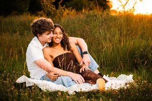 Couple sitting on blanket in golden hour field, embracing and smiling warmly during romantic outdoor picnic.