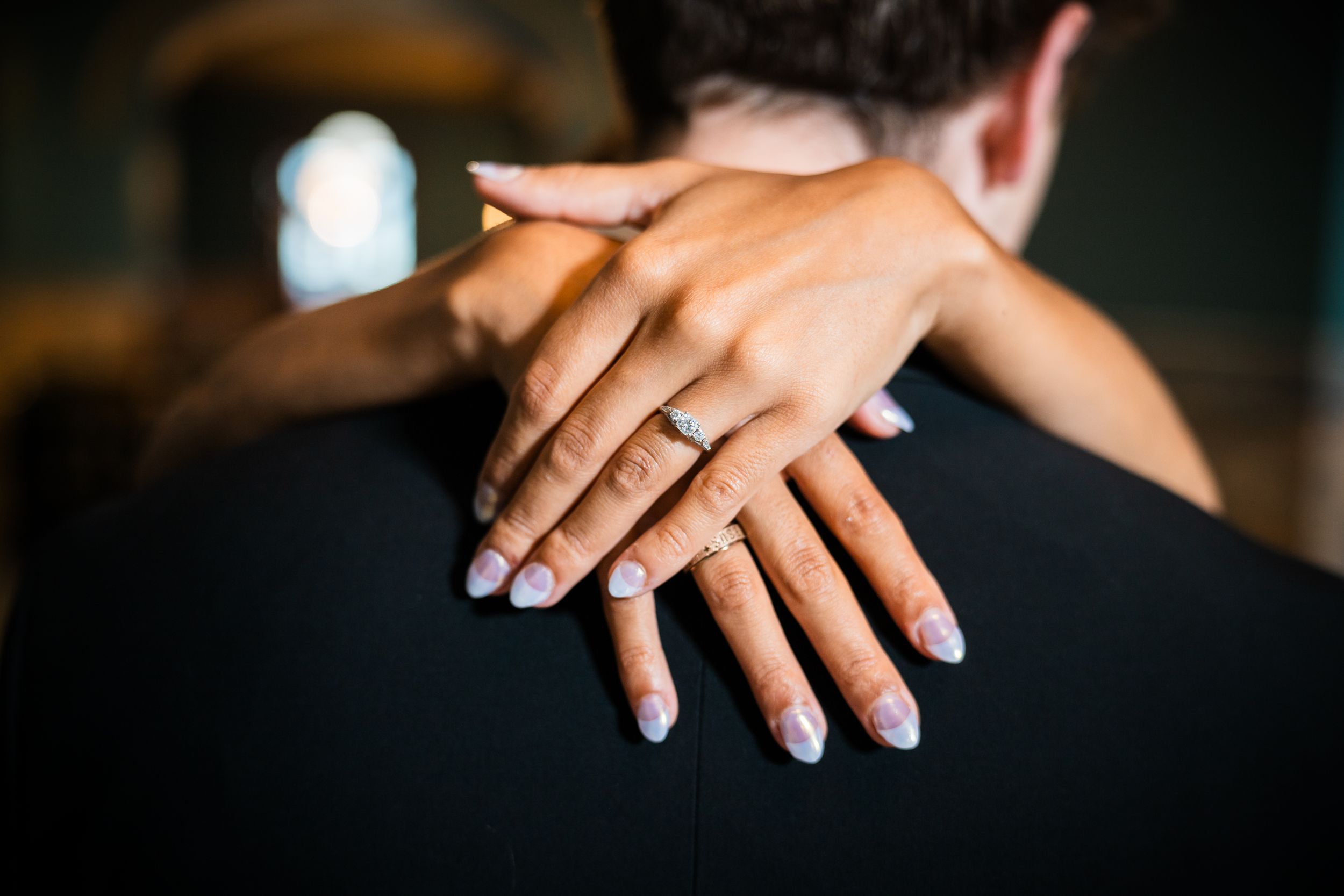 Close-up of a woman's hand with an engagement ring and manicured nails resting on a man's shoulder.