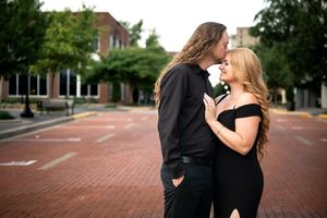 Couple in matching black outfits sharing a romantic moment on a brick plaza, surrounded by lush green trees.