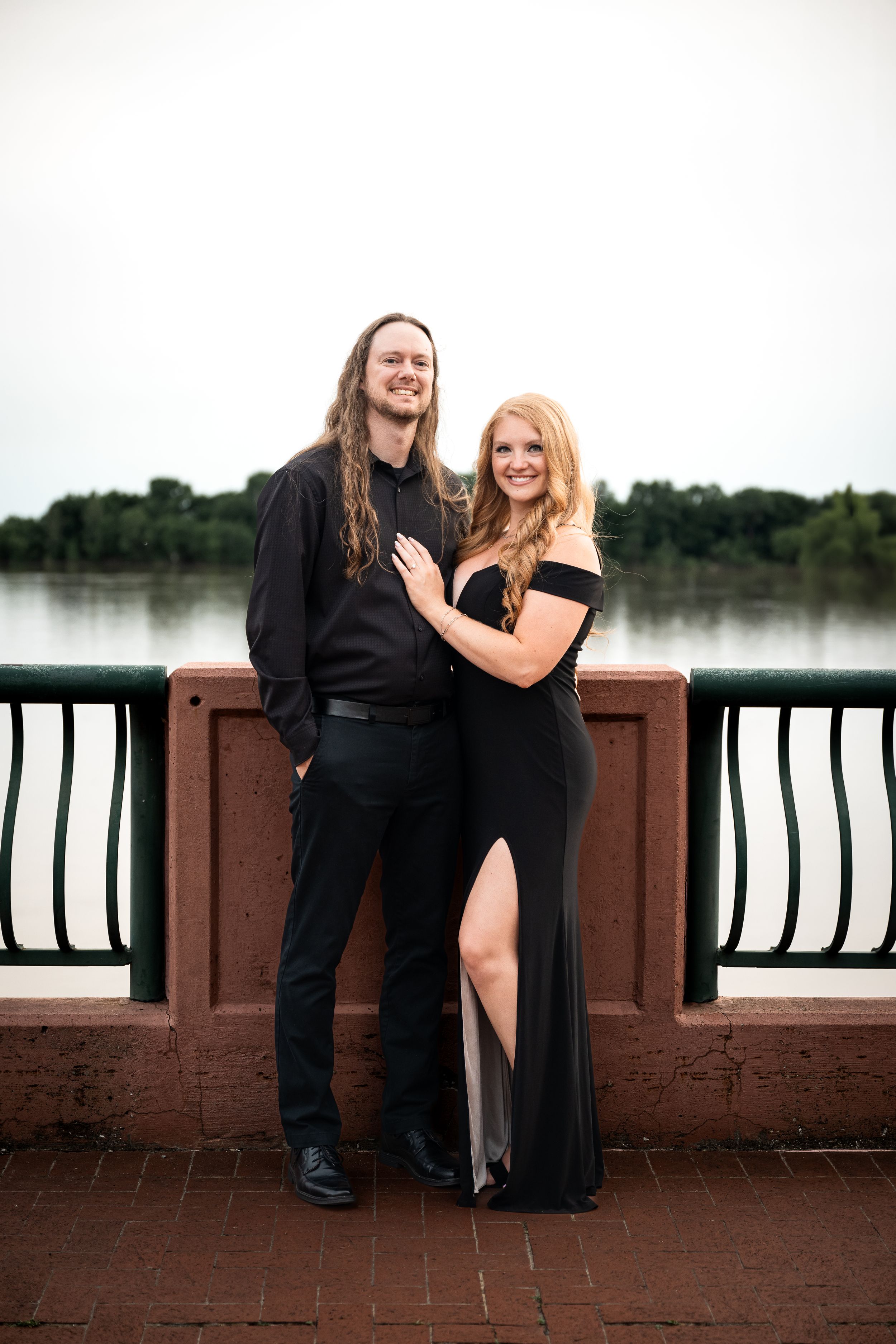 Couple in matching black formal attire posing on a bridge with a river and trees in the background.