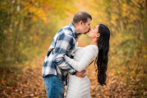 Couple sharing a romantic kiss on a tree-lined autumn path, surrounded by vibrant yellow and orange fall foliage.
