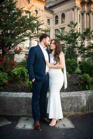 Couple in white dress and navy suit share romantic moment outdoors near historic building with lush greenery.