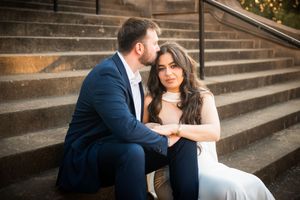 Couple sitting on stone steps, man in navy suit kissing woman's head, she holds his hands wearing white dress.