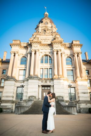 Couple sharing a romantic kiss in front of a grand neoclassical building with columns under a clear blue sky.