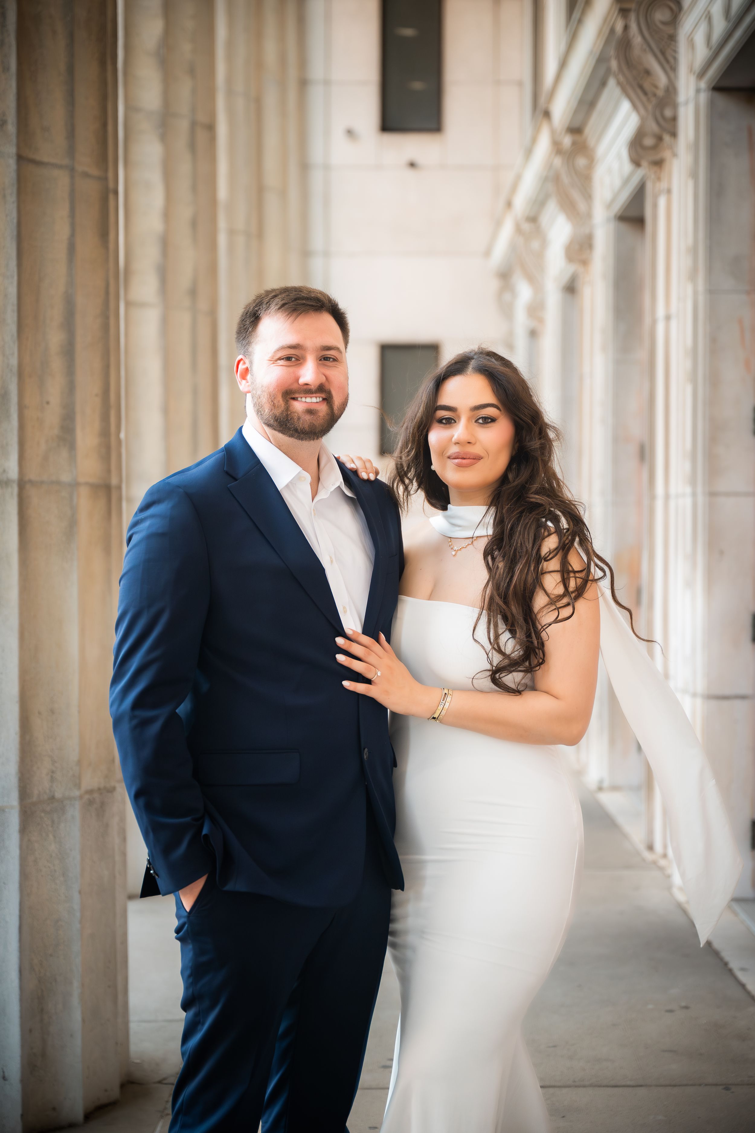 Couple posing elegantly outdoors, man in navy suit and woman in white fitted dress, smiling beside classical stone columns.