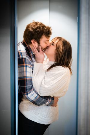 A couple sharing a romantic kiss in an elevator, man in plaid shirt and woman in white sweater embracing tenderly.