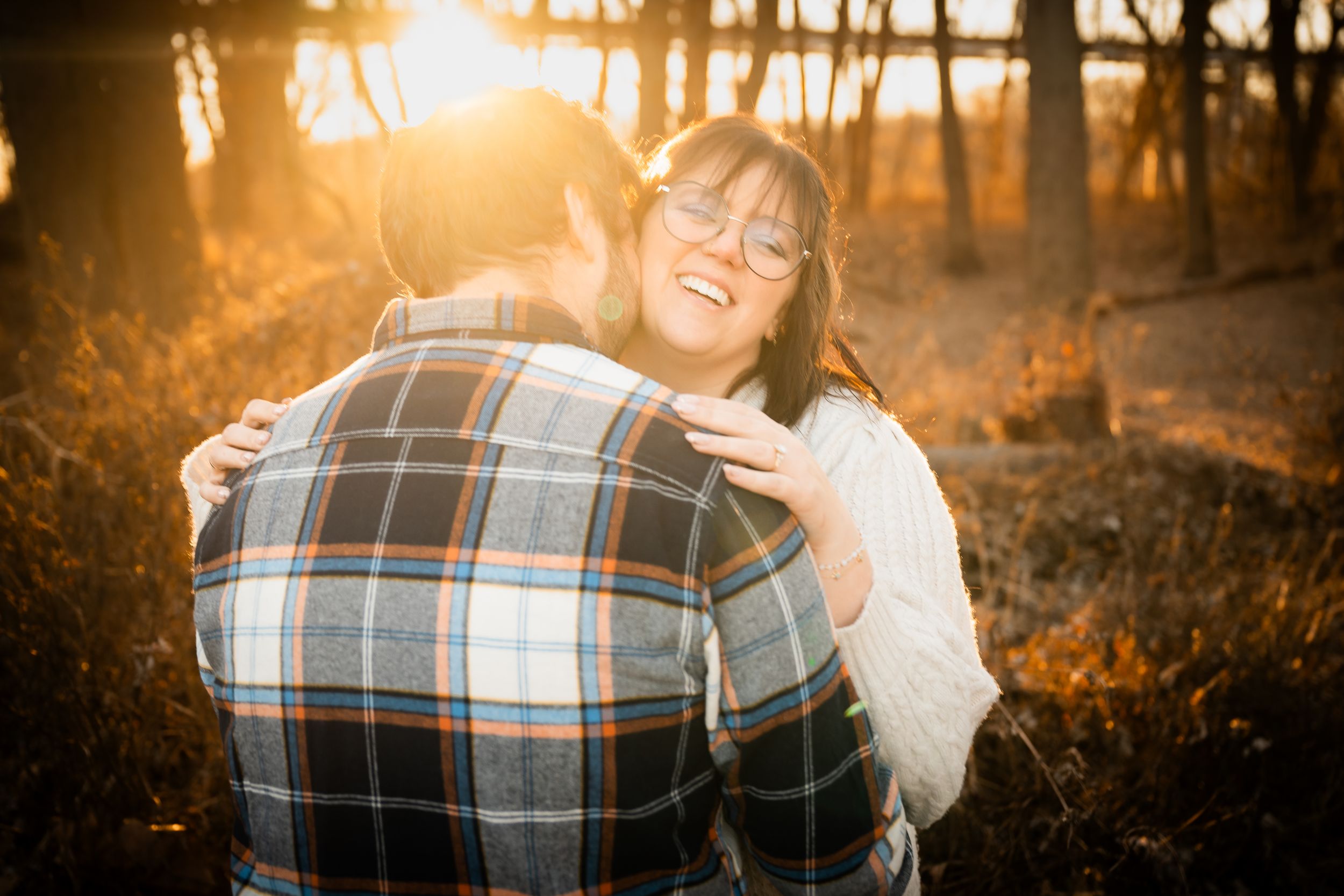 A joyful woman in glasses embraces a man in a plaid shirt during a golden sunset in an autumn forest.