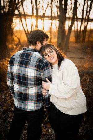 Couple embracing at golden hour in autumn woods, woman in white sweater smiling, man in plaid shirt.