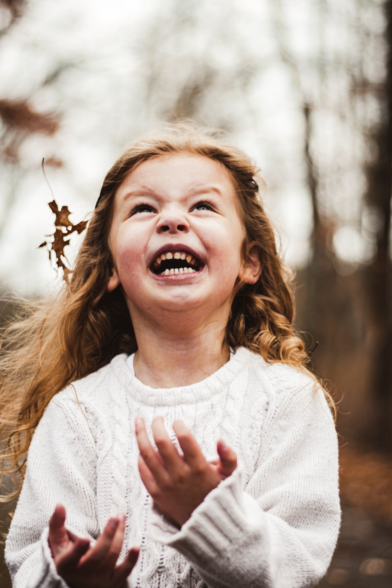 Young child in white sweater laughing joyfully with curly hair blowing in wind during an autumn day.