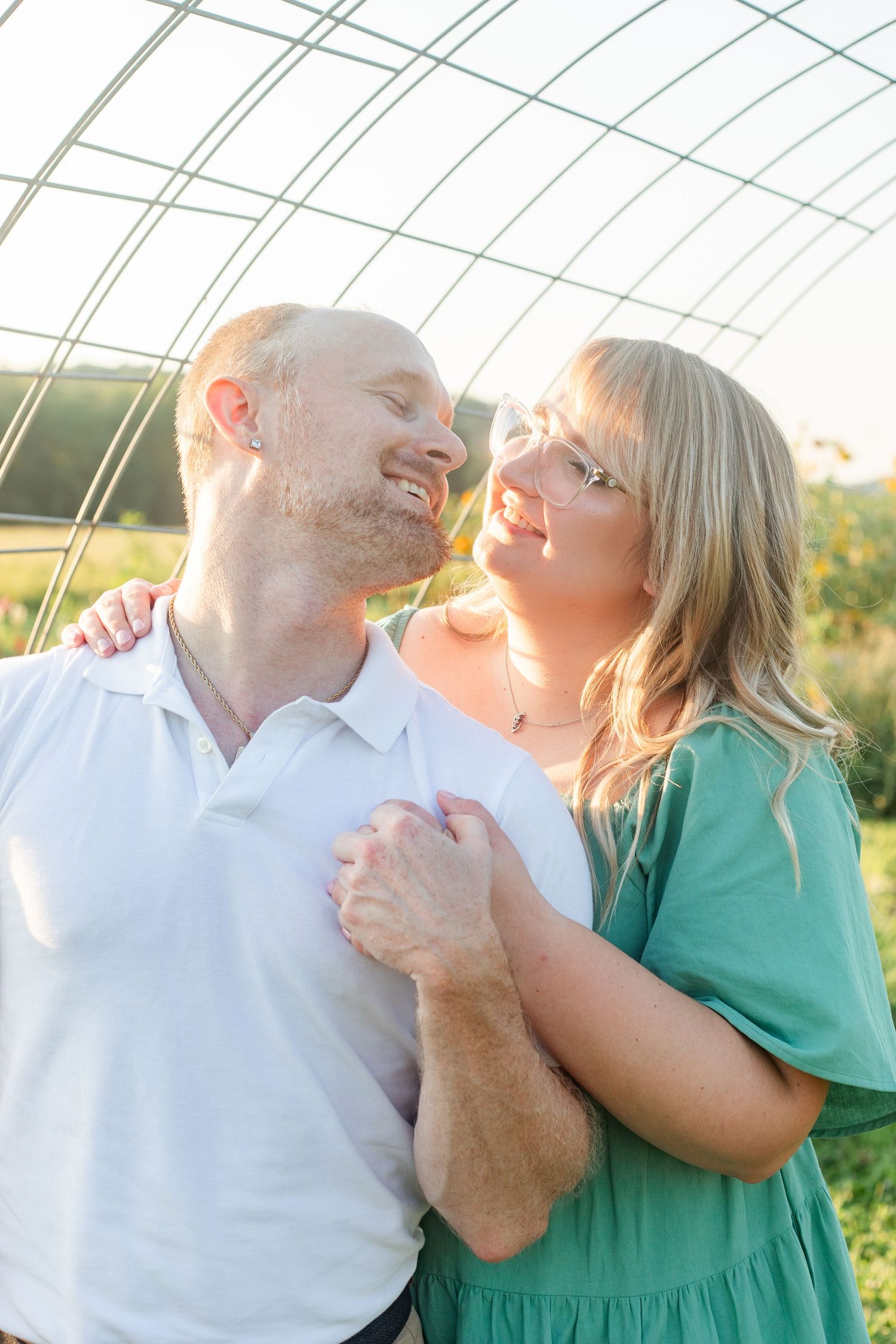 A couple shares a tender moment together in warm sunlight under a greenhouse structure.