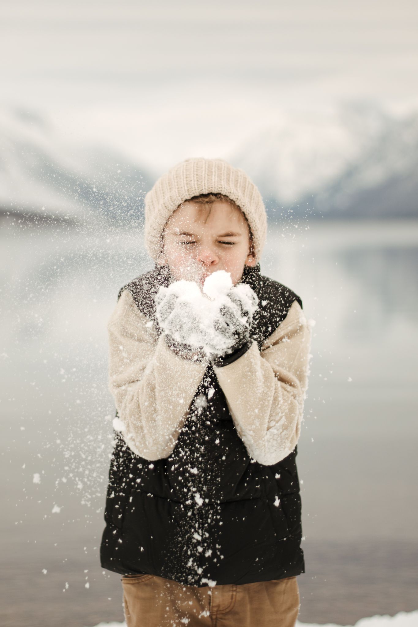 Person in winter attire blowing snow from mittened hands near a misty mountain lake.