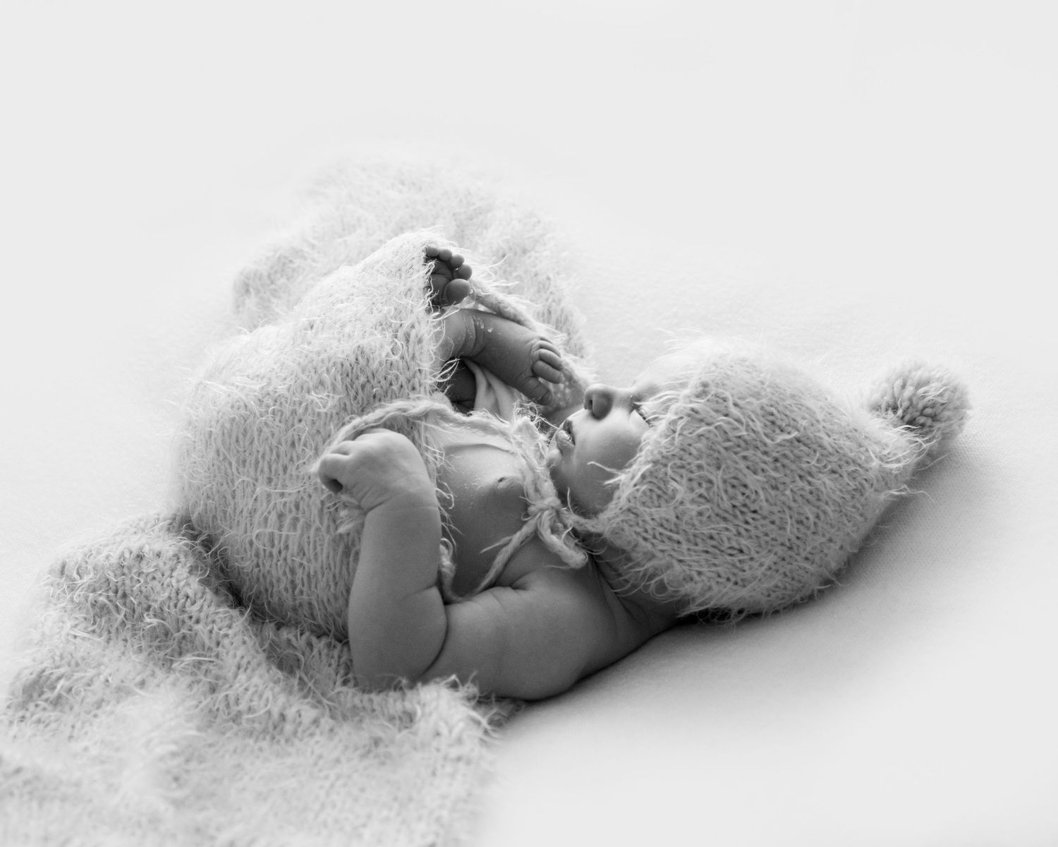 A black and white image of a sleeping baby. The baby is content and wearing a fluffy hat and wrapped comfortably in a blanket.