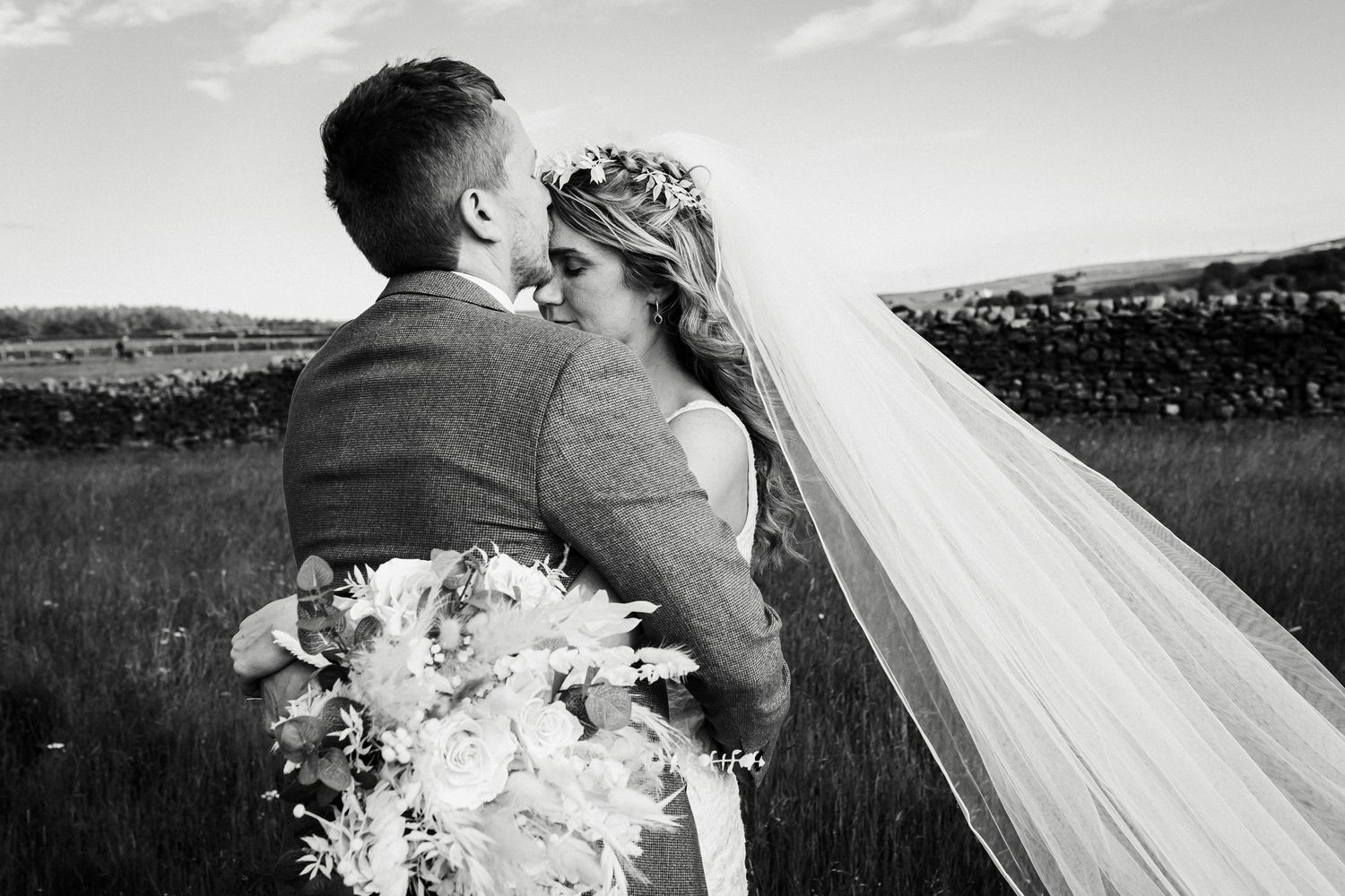 A romantic black and white wedding photo showing a couple embracing in a field with a white bouquet.