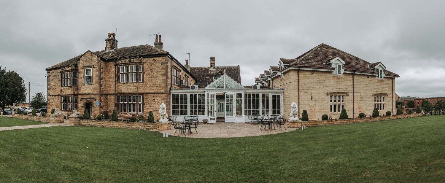 Panoramic view of an elegant stone mansion with conservatory and manicured lawn.