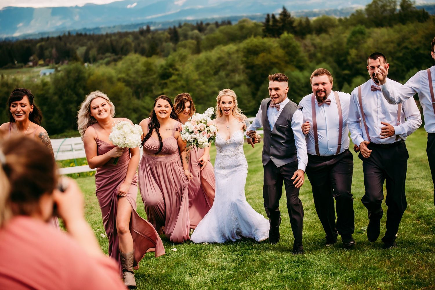 Wedding party in pink and grey attire walking together on grassy hillside.