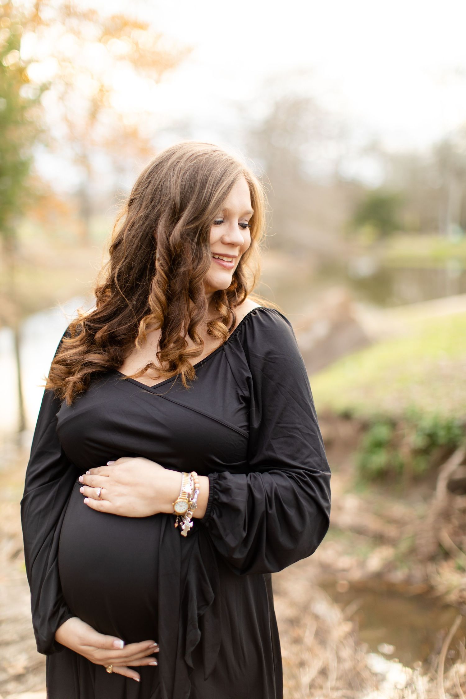 Woman in black dress poses for maternity photo in natural outdoor setting by stream at sunset.
