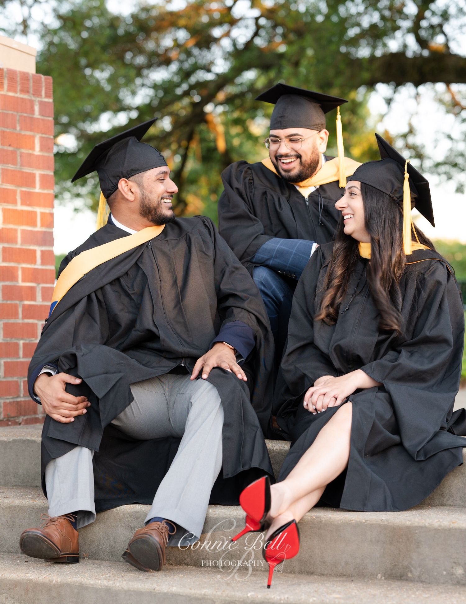 Three graduates in black caps and gowns share a candid moment on steps.