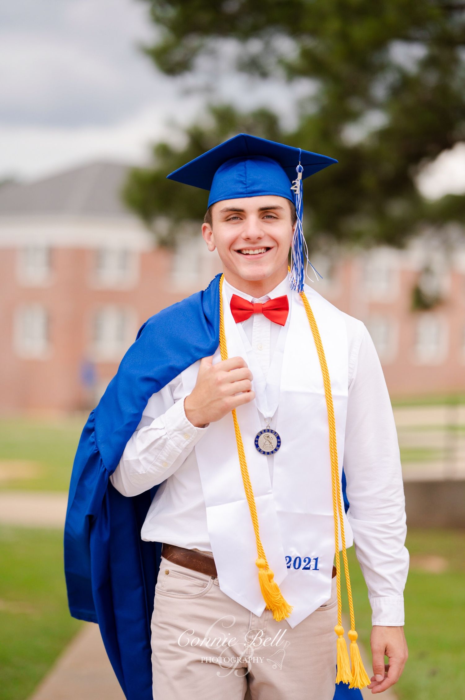 Graduate in blue cap and gown with red bowtie and gold cords poses outdoors on campus.