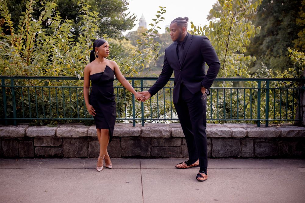 A couple in elegant black attire holds hands while standing on a stone terrace.