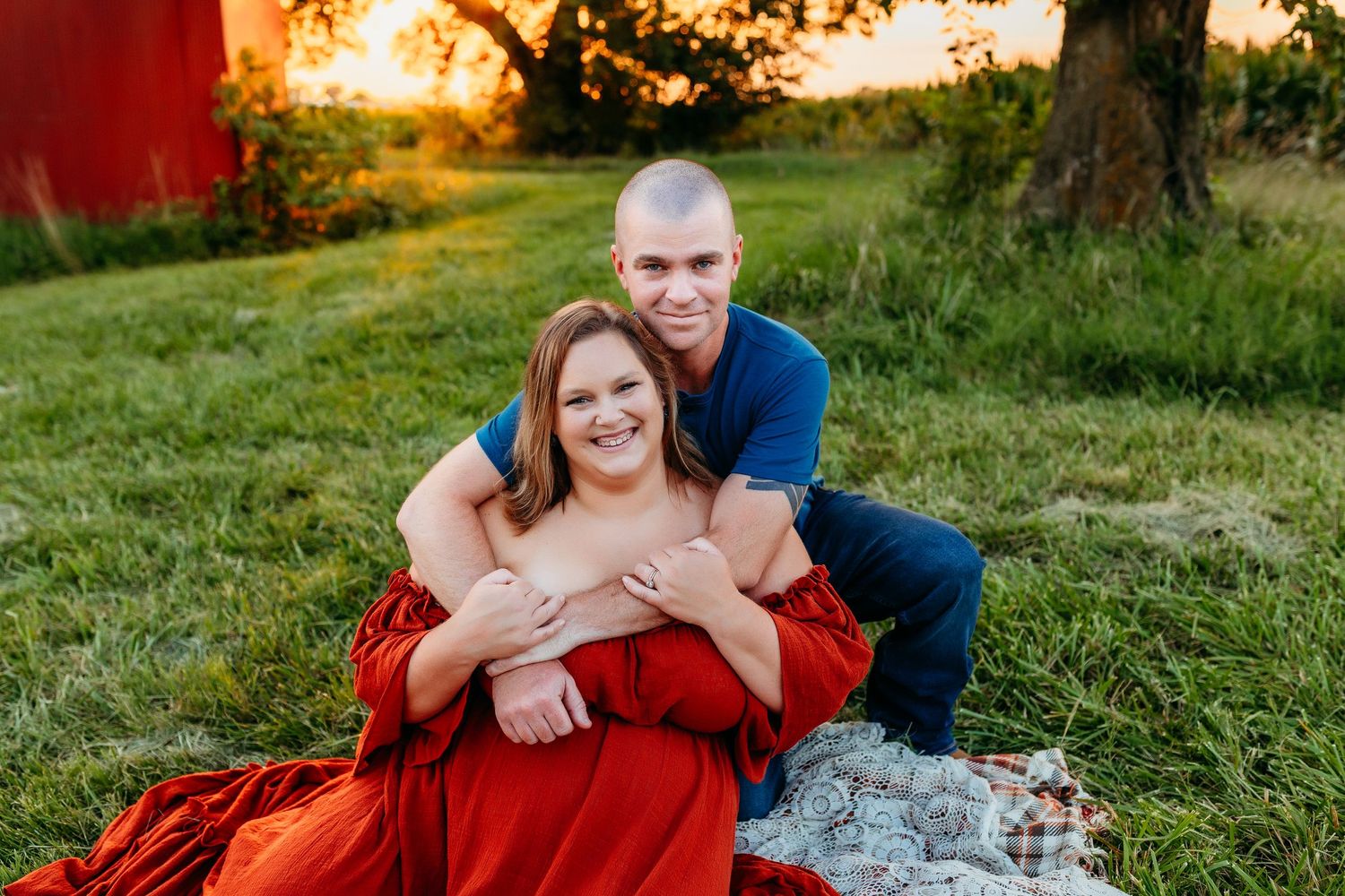 A romantic outdoor photoshoot on a blanket near a red barn at sunset.