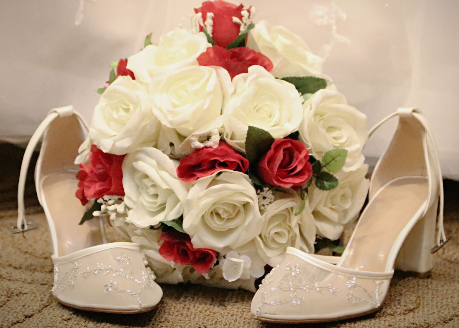 A bridal bouquet of red and white roses sits between sparkly wedding shoes.
