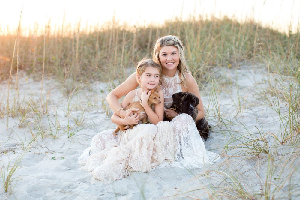 Two people sitting in sand dunes with chihuahua and black dog at sunset.