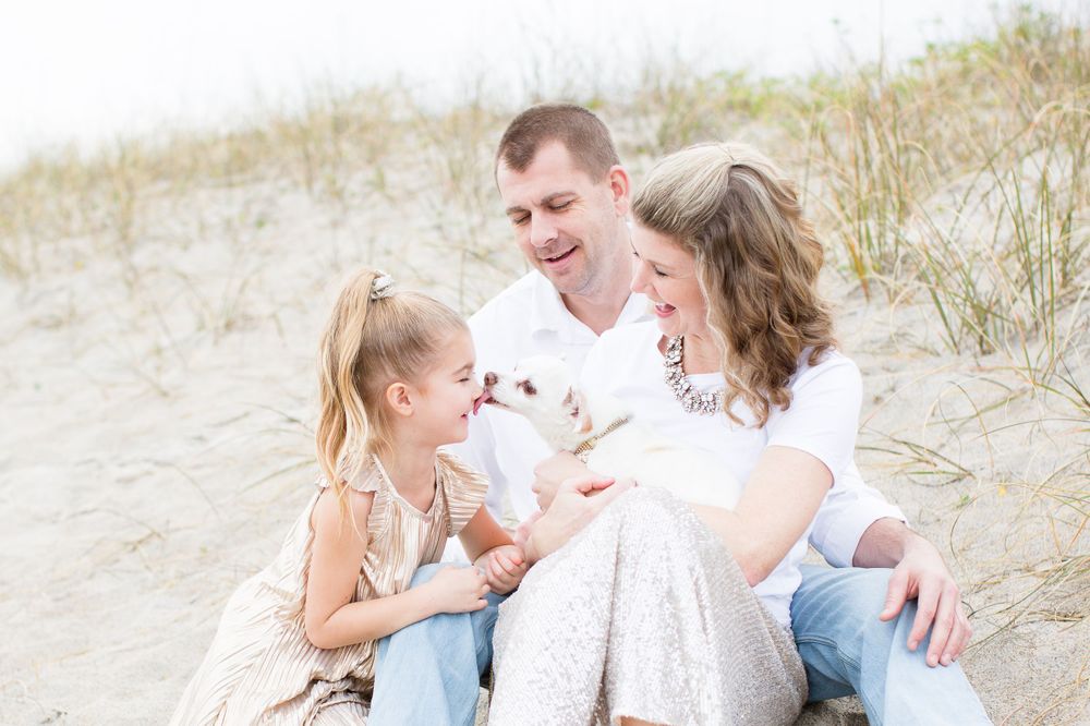 Family sitting together on sandy beach dunes with small white dog.