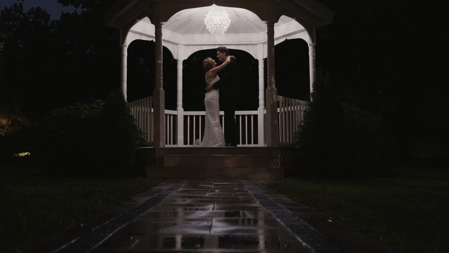 A silhouette in a white gazebo at night with rain-covered ground.
