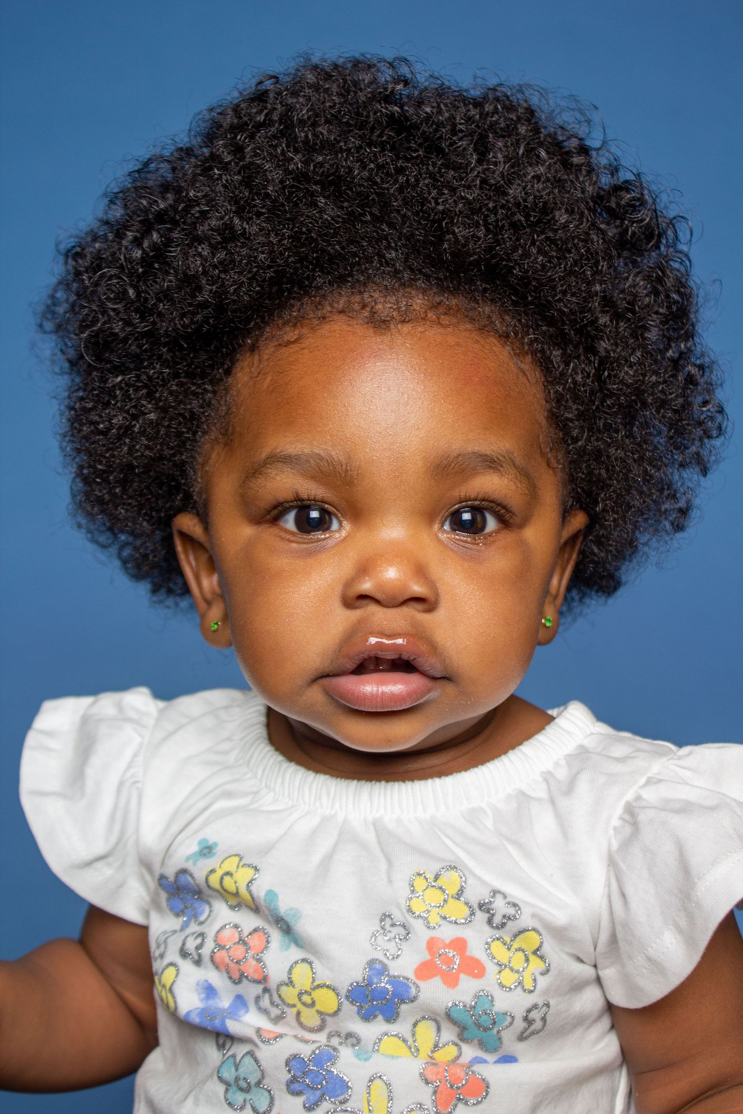 Adorable baby girl with curly afro hair wearing floral dress poses for portrait photo.