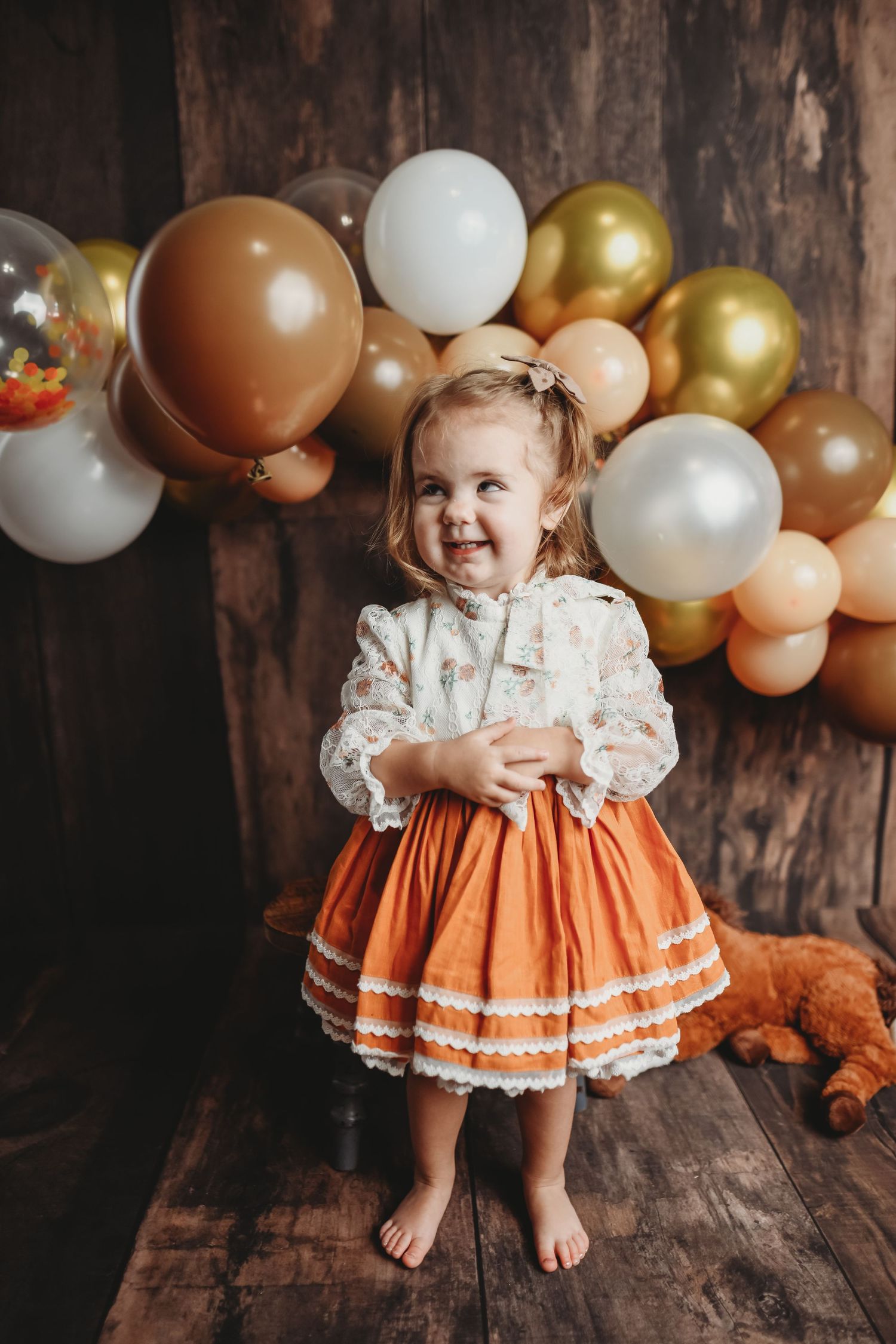 Child in orange dress sitting with gold and white balloon arch backdrop.