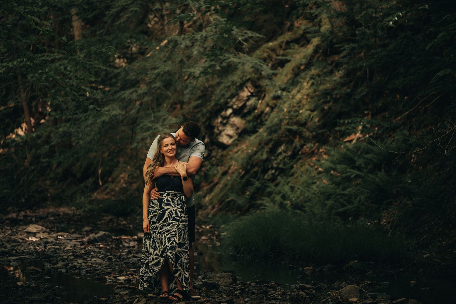 A couple embraces in a lush forest setting with dramatic lighting and greenery.