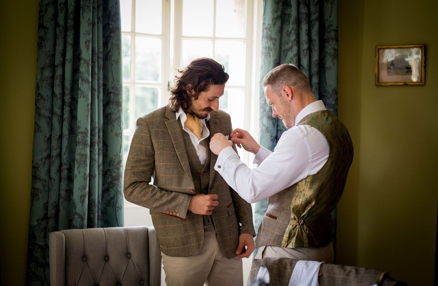 Two people in formal wear assist with adjusting a boutonniere in an elegant room with green curtains.