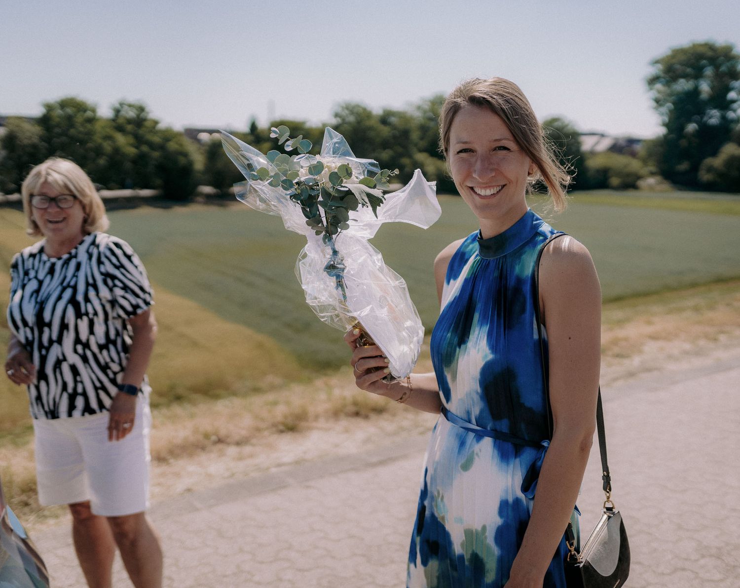Woman in blue tie-dye dress holds white floral bouquet outdoors on sunny day.
