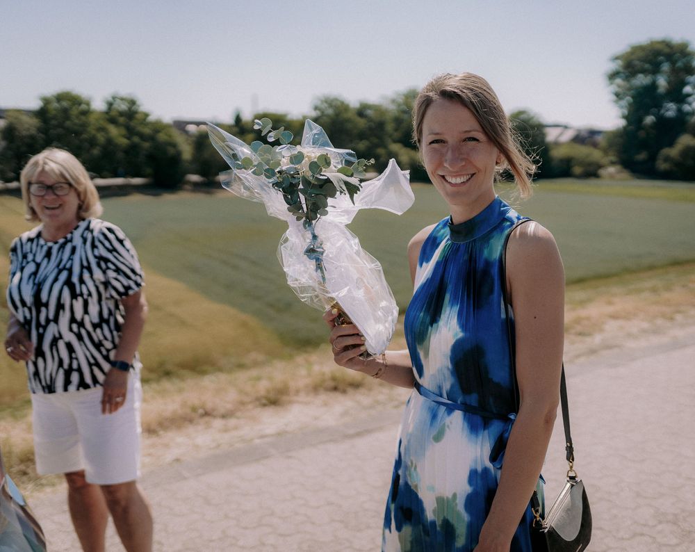 Woman in blue tie-dye dress holds white floral bouquet outdoors on sunny day.